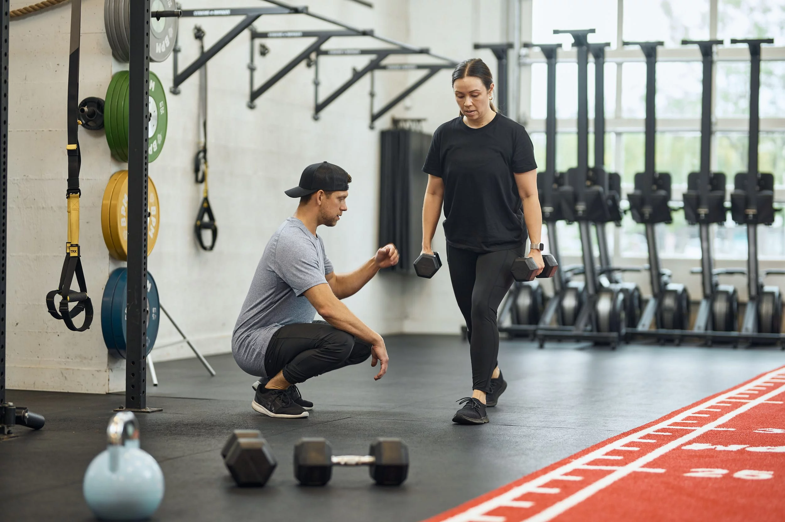 A personal trainer assists a woman with dumbbell exercises in a gym setting, showcasing fitness training and strength building.